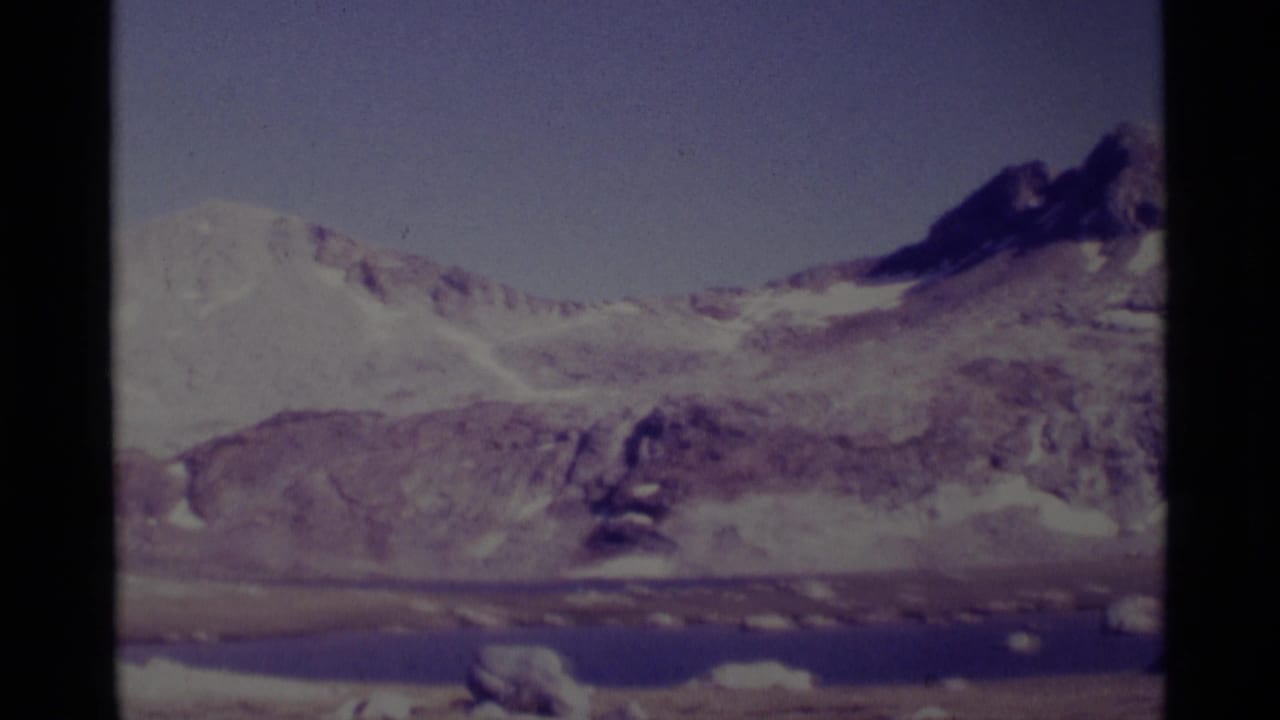 Minaret Wilderness landscape with a darker mountain peak, snowfields, rocky terrain, and lake water visible in a vintage 8mm frame.
