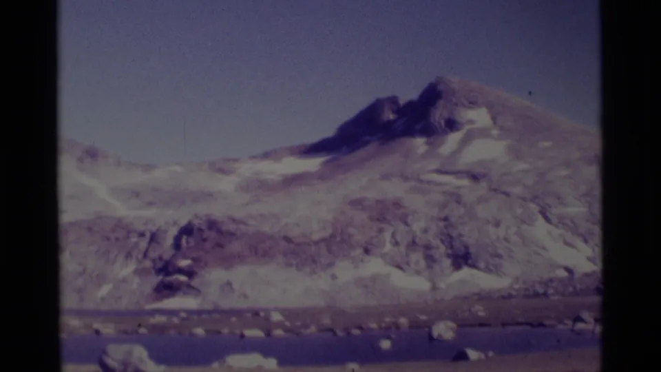 1975 home-movie view of a snow-streaked mountain peak in the Minaret Wilderness area, with lake water and loose rocks below.