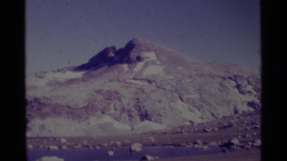 Wide 8mm view of the Minaret Wilderness area with snow patches on rugged mountains, a calm lake, scattered rocks, and visible frame borders.