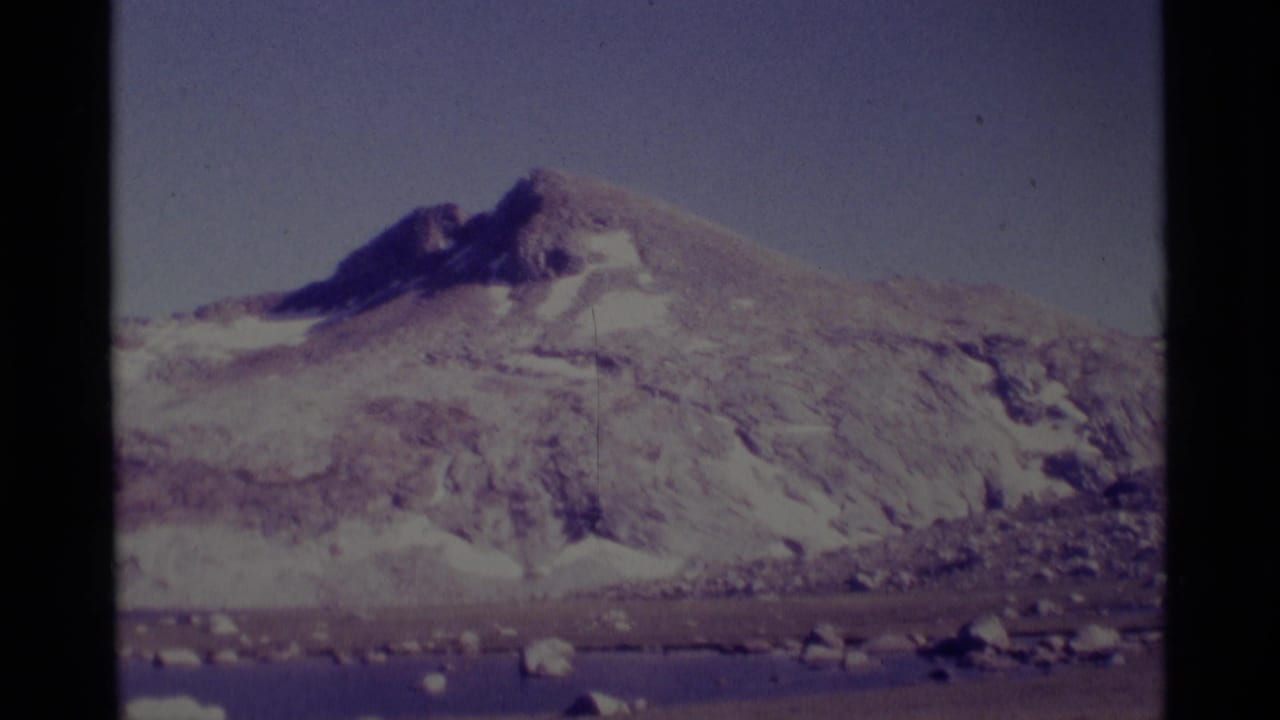 1975 8mm view of a snow-streaked mountain peak in the Minaret Wilderness area, framed by rocky foreground and dark film gate edges.