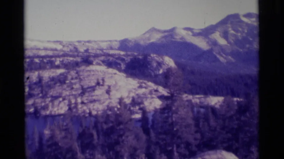 Faded 8mm frame of rocky high-country foreground, pine forest, and distant snow-streaked ridges in California wilderness metadata context.