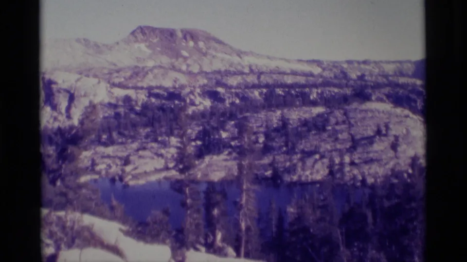 Wide 1975 8mm landscape view of the Minaret Wilderness area with lake water, snowy slopes, forest, and rocky Sierra terrain.