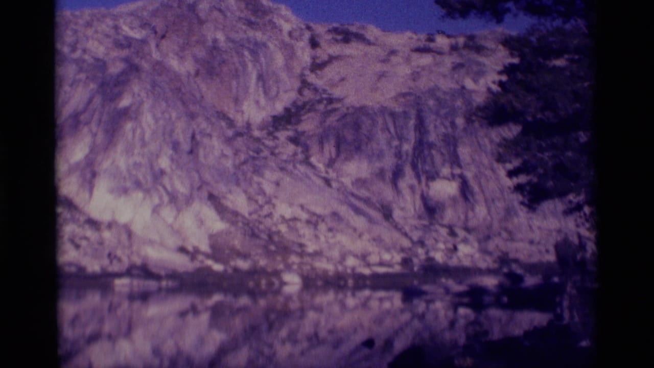 1975 8mm view of a rocky Minaret Wilderness cliff reflected in still water, with dark tree silhouettes at the frame edge.