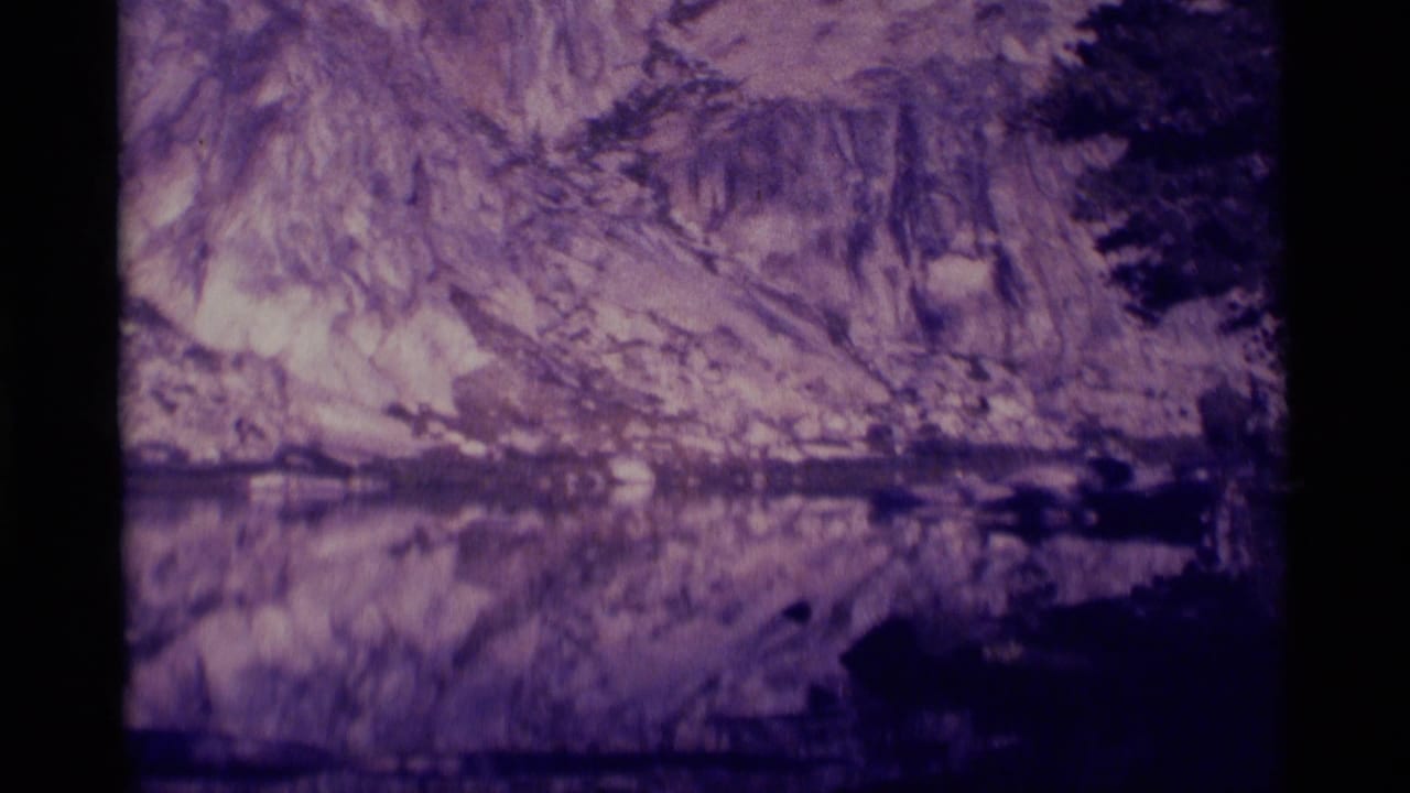 Closer view of a rugged cliff and its reflection across still water, framed by dark trees.
