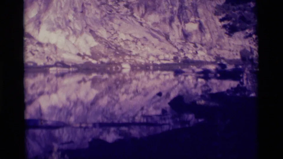 Final 1975 Minaret Wilderness frame holding on the cliff and mirrored water, with dark trees and aged blue-purple 8mm color.