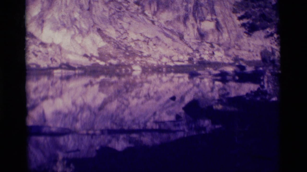 1975 Minaret Wilderness home-movie frame showing a mountain lake reflecting rocky cliffs and a shaded shore.