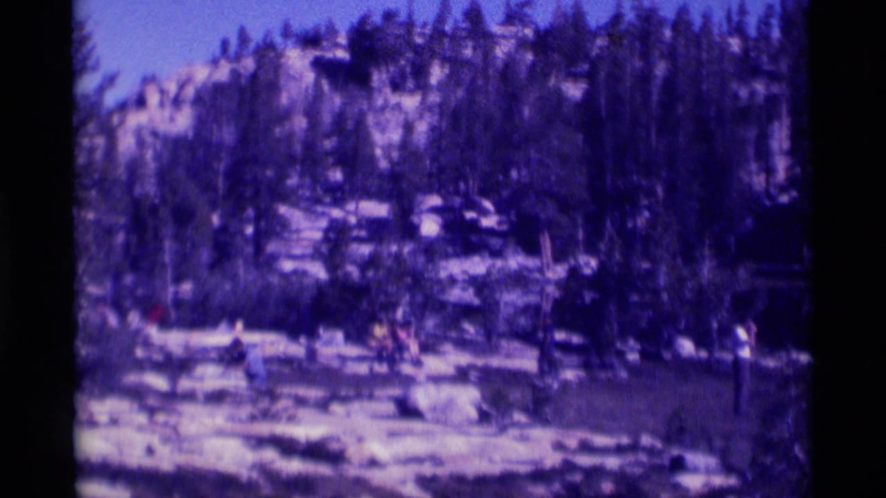 1975 Minaret Wilderness frame with distant people among rocks and evergreen trees beside a mountain lake.