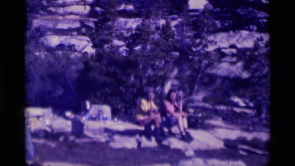 1975 Minaret Wilderness frame showing distant people seated or standing on lakeside rocks with trees behind them.
