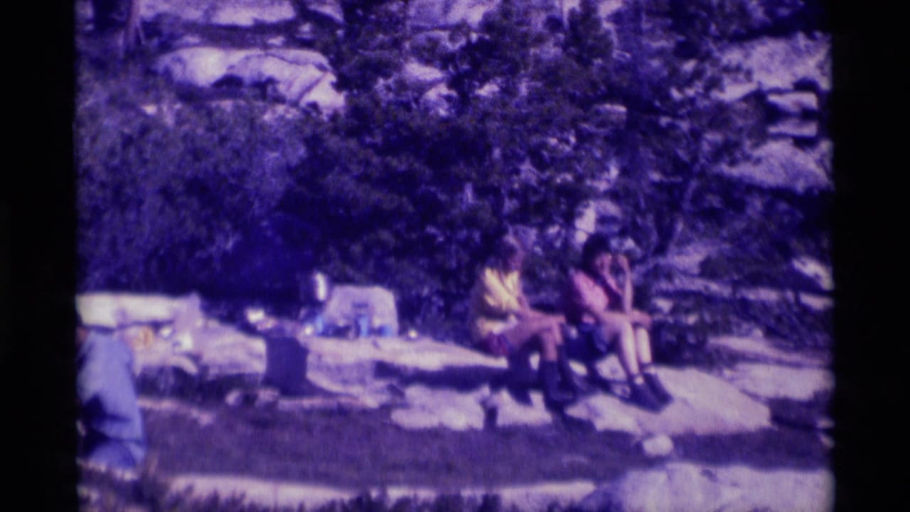 Two people sit on a broad rock near picnic items in a wooded rocky setting.
