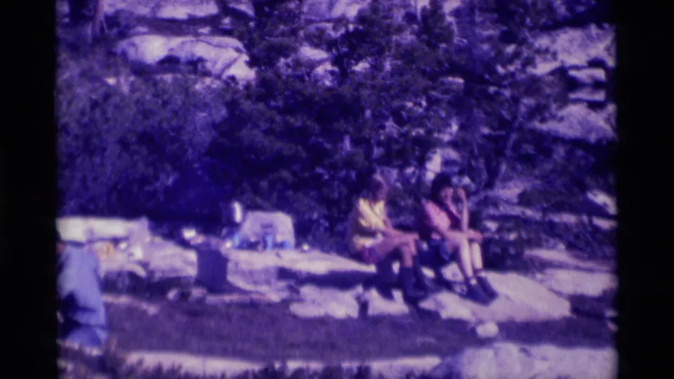 Two visible people sit on a large rock during a Minaret Wilderness picnic, surrounded by dark trees, stone, and soft 8mm color shift.