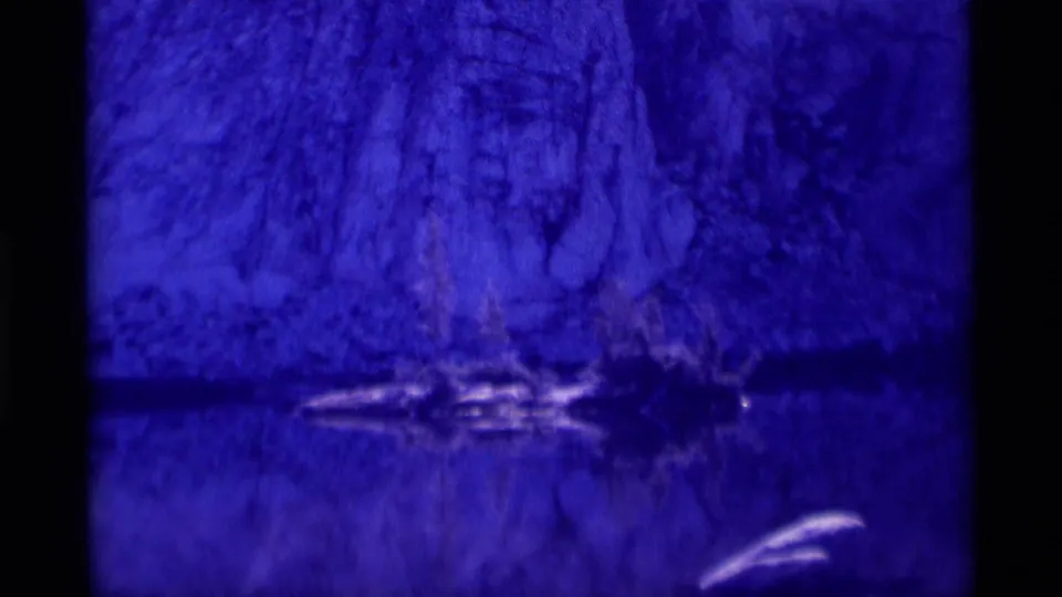 Heavily tinted 1975 wilderness frame showing a soft cliff reflection in water, with 8mm grain and a nearly monochrome blue cast.
