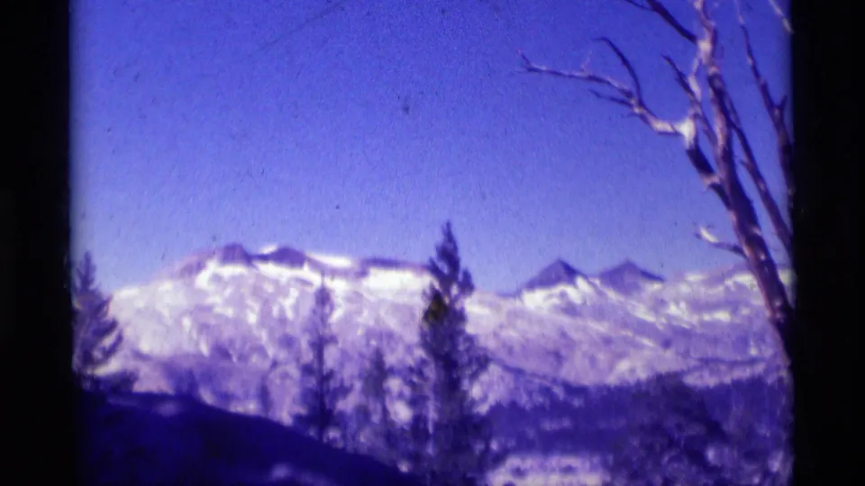 1975 8mm mountain view in the Minaret Wilderness area, with snowy ridges, evergreens, a bare tree, and blue-purple sky.