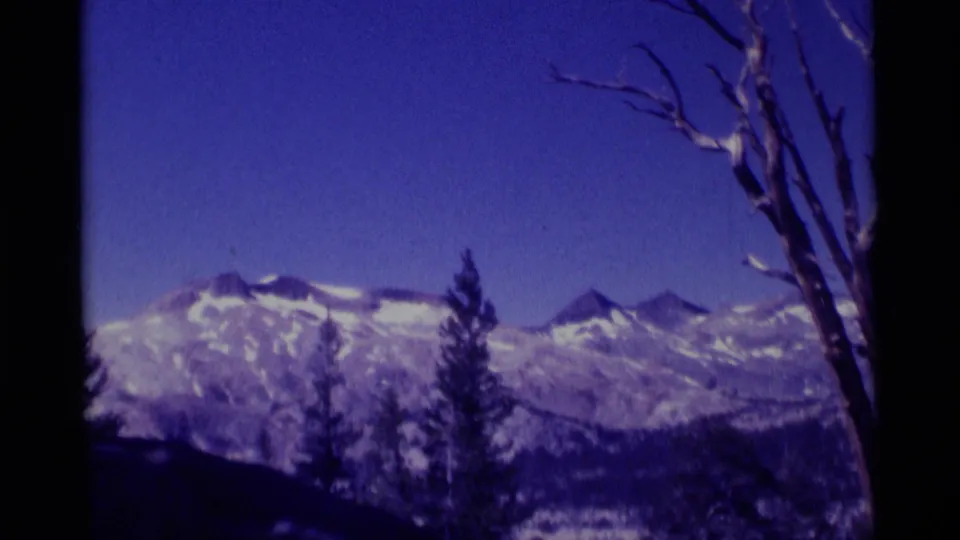 Vintage 8mm view of Minaret Wilderness mountain ridges, snow fields, dark tree silhouettes, and a bare trunk at frame edge.
