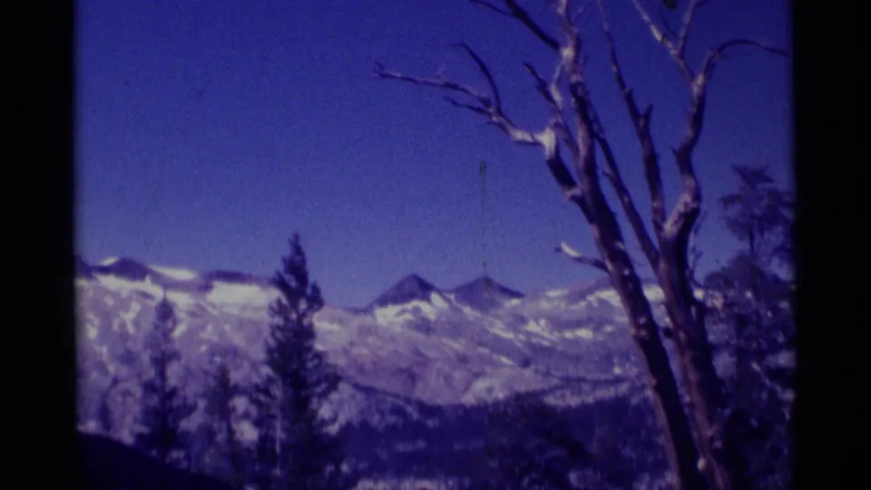 Minaret Wilderness mountain view on 8mm film, showing snow-dusted peaks, evergreens, and a leafless tree against a blue sky.