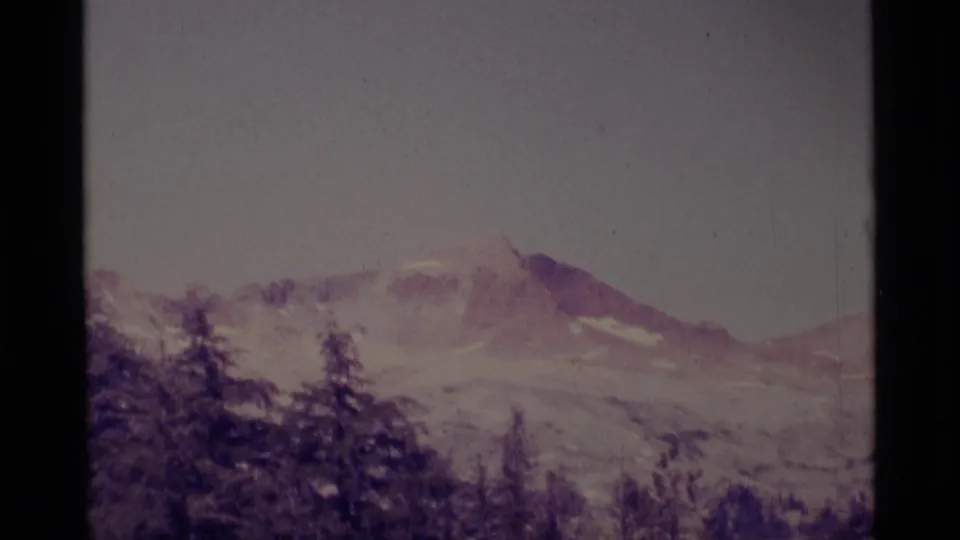 1975 8mm view of a mountain summit in the Minaret Wilderness area, seen beyond dark forest with a faded purple film cast.