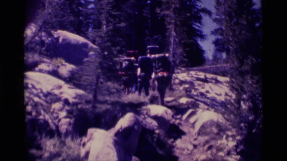 Distant hikers with backpacks move along a rocky wooded trail in 1975 8mm Minaret Wilderness home-movie footage.