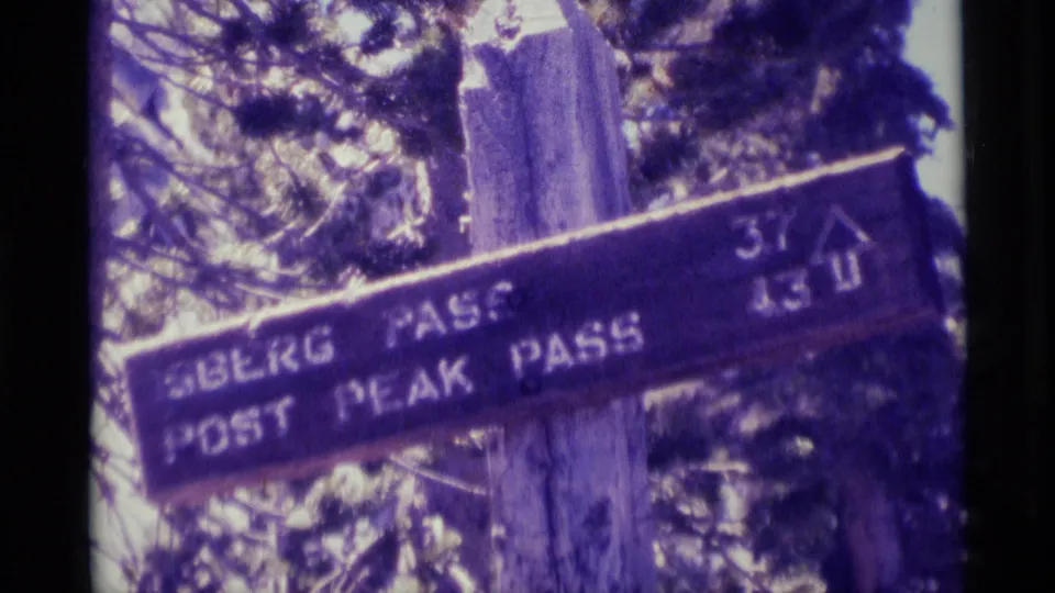 A wooden wilderness trail sign for Sberg Pass and Post Peak Pass appears among trees in the 1975 8mm clip.
