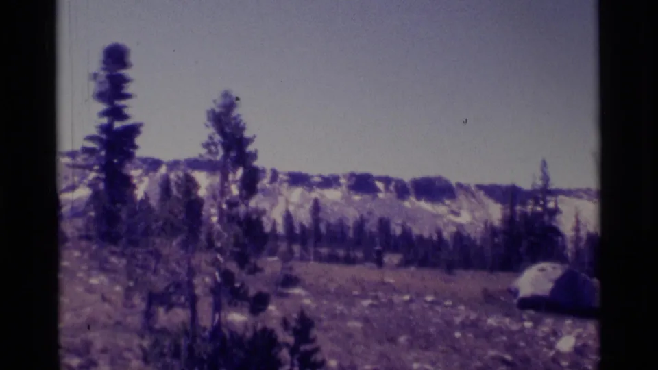 1975 8mm landscape view of Minaret Wilderness with pines, a snow-streaked ridge, rocks, and pale sky.