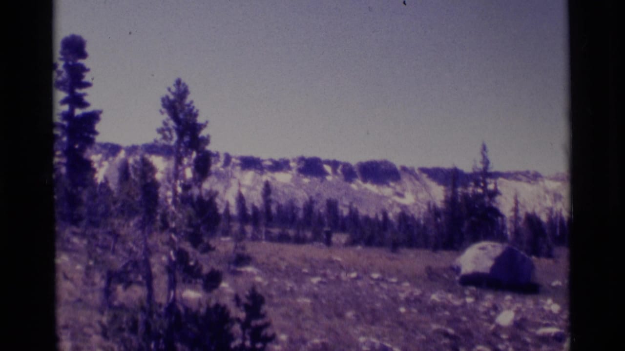 1975 8mm view of Minaret Wilderness terrain with scattered pines, meadow slope, boulders, and snow-covered mountains beyond.
