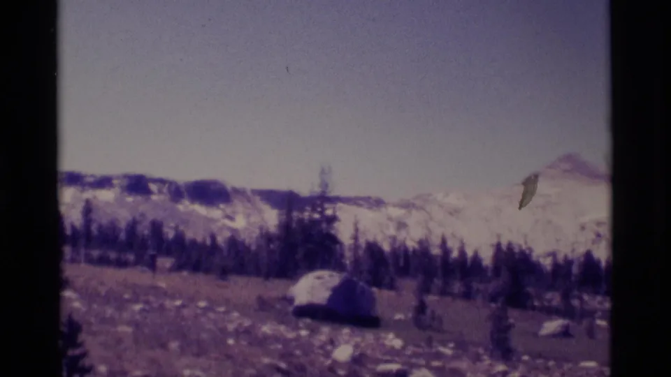 1975 8mm mountain landscape in Minaret Wilderness, with a pine line, meadow rocks, a large boulder, and snowy peaks under clear sky.