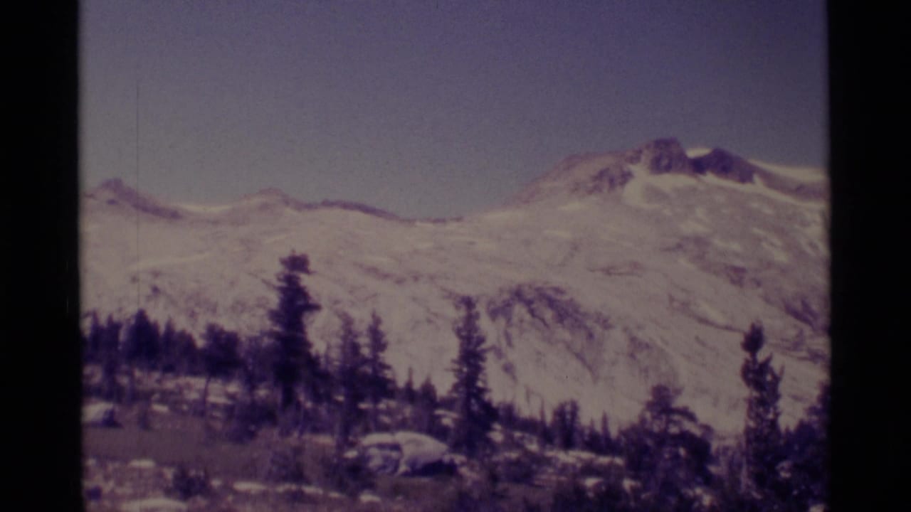 1975 8mm view of snow-dusted mountains in Minaret Wilderness, California, framed by evergreen trees and dark film edges.
