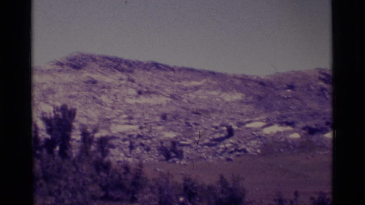 1975 8mm view of the Minaret Wilderness area showing snow-dappled foothills, pale sky, flat valley land, and dark trees at frame edge.