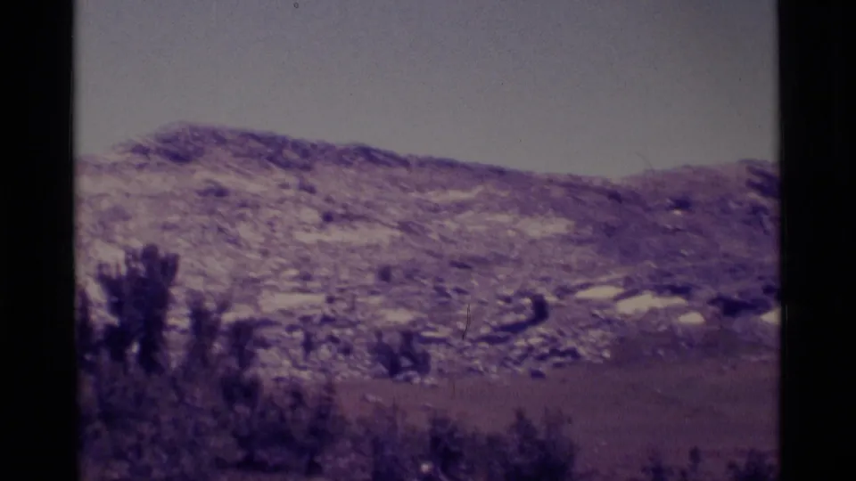 1975 8mm view of the Minaret Wilderness area showing snow-dappled foothills, pale sky, flat valley land, and dark trees at frame edge.