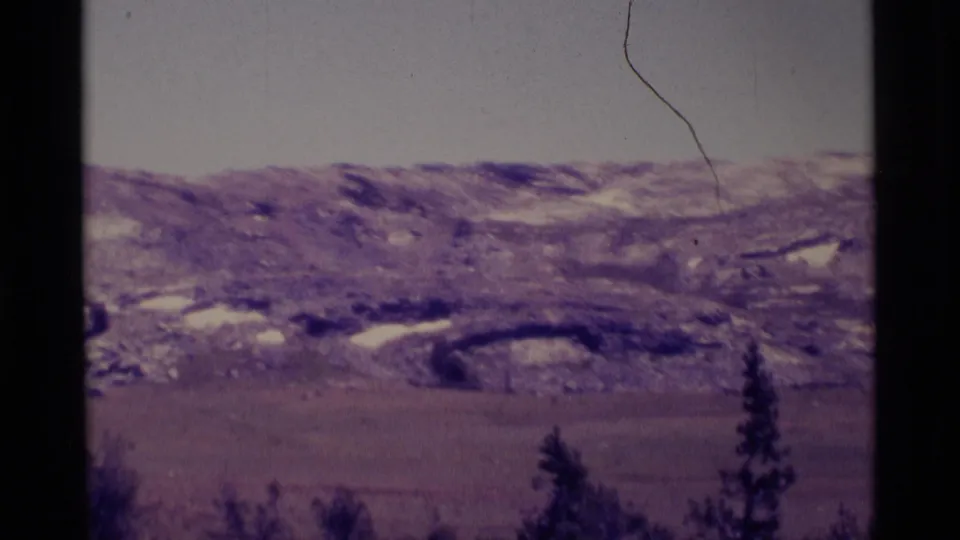 1975 home-movie frame from the Minaret Wilderness area showing rocky snow patches, flat valley ground, evergreen tops, and visible film scratches.