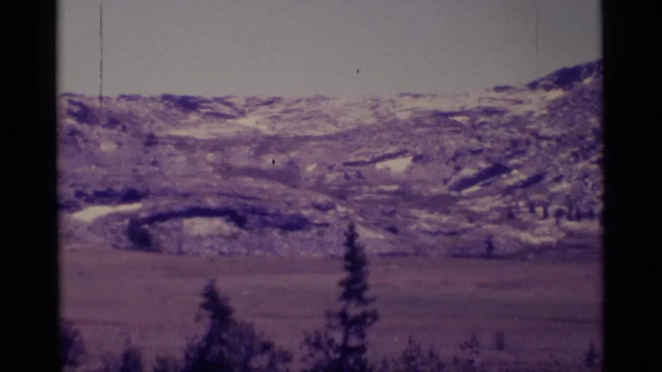 Archival 8mm view of the Minaret Wilderness area with a pale sky, rocky snow patches, open valley land, and dark evergreen foreground trees.