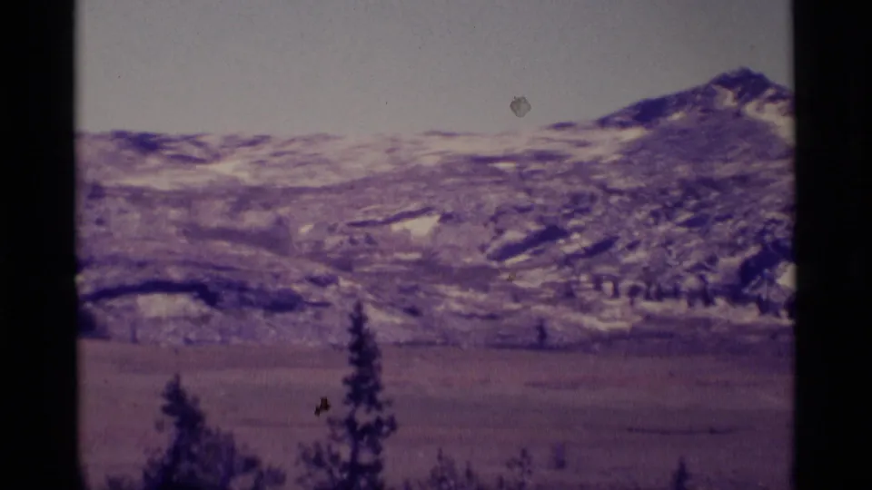 1975 8mm view of Minaret Wilderness mountains, with snow patches on ridges above a broad meadow and conifer silhouettes.