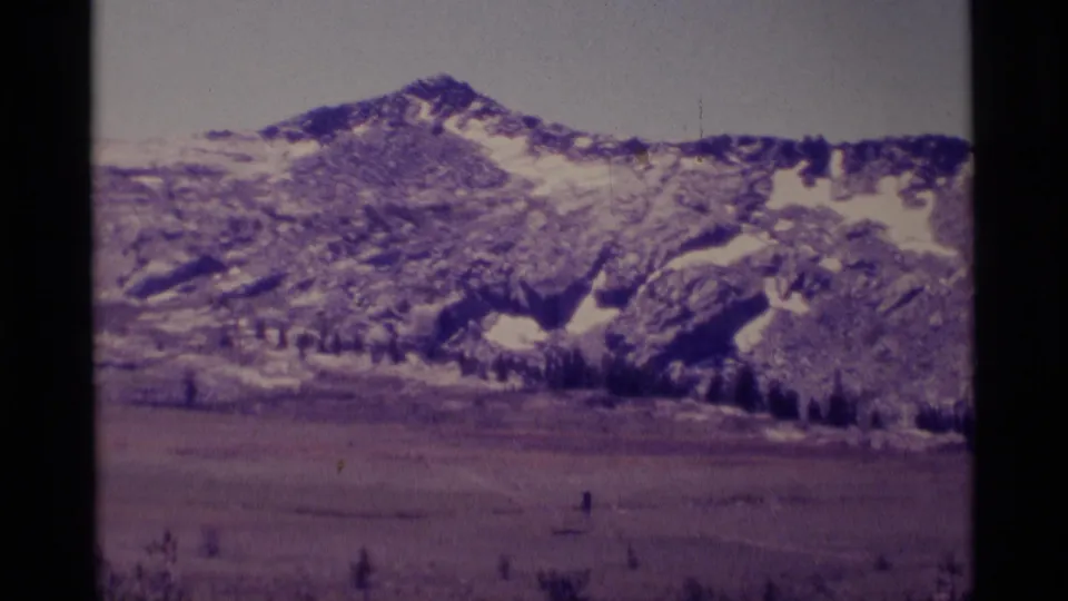 1975 home-movie frame of Minaret Wilderness, with a snow-streaked ridge, open meadow, and dark conifers near the base.