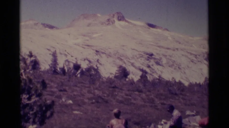 Distant hikers in 1975 Minaret Wilderness footage rest on a hillside below a broad snow-covered mountain slope.