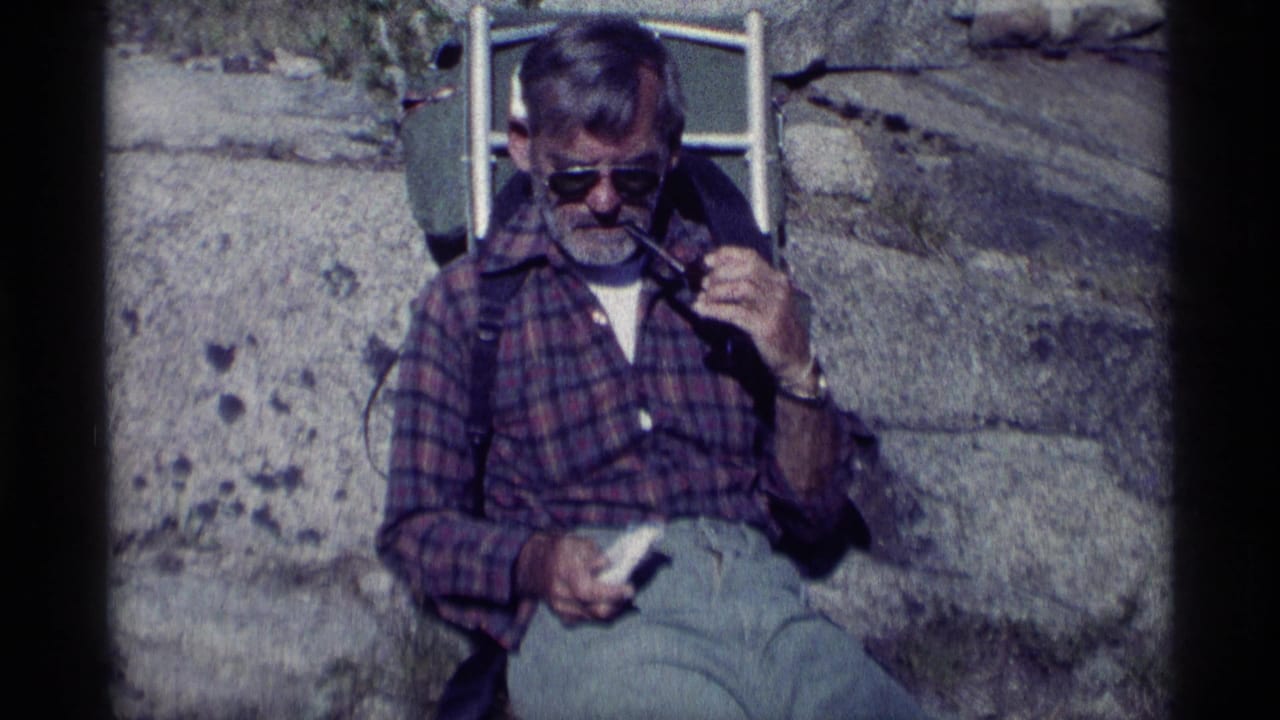 Adult man in plaid shirt and sunglasses sits against a rock in Minaret Wilderness, raising his pipe during a hiking break.