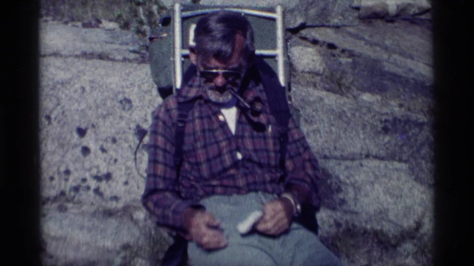 In 1975 California wilderness footage, a seated hiker smokes a pipe with a metal-frame pack visible behind him.