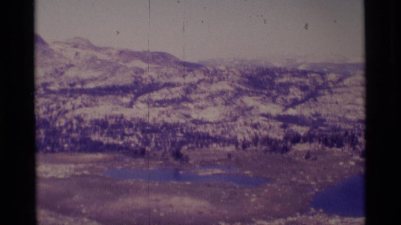 1975 8mm Minaret Wilderness panorama showing snow-streaked mountains, dark tree cover, open meadow, and blue alpine lakes.