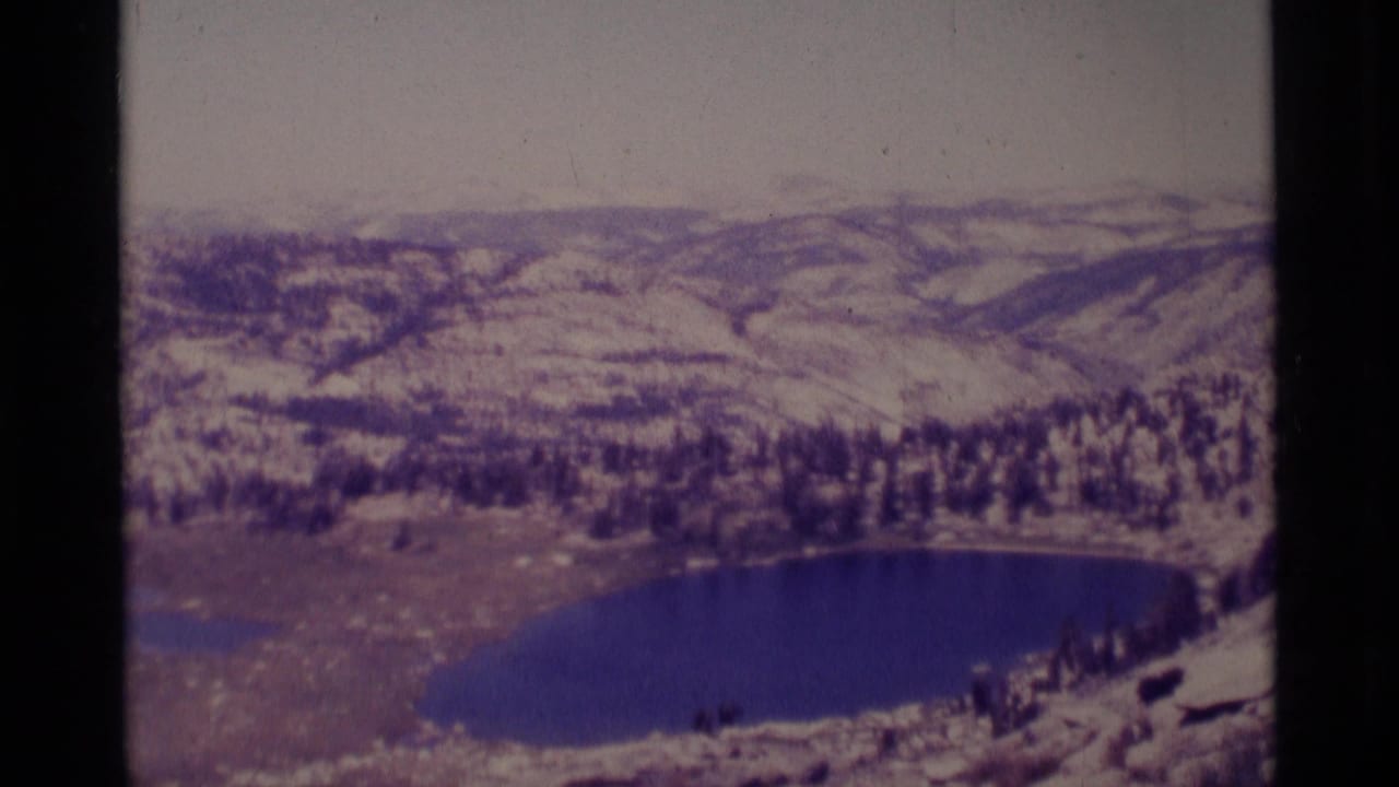 1975 8mm view of a Minaret Wilderness lake surrounded by evergreen trees, pale slopes, and distant ridgelines.