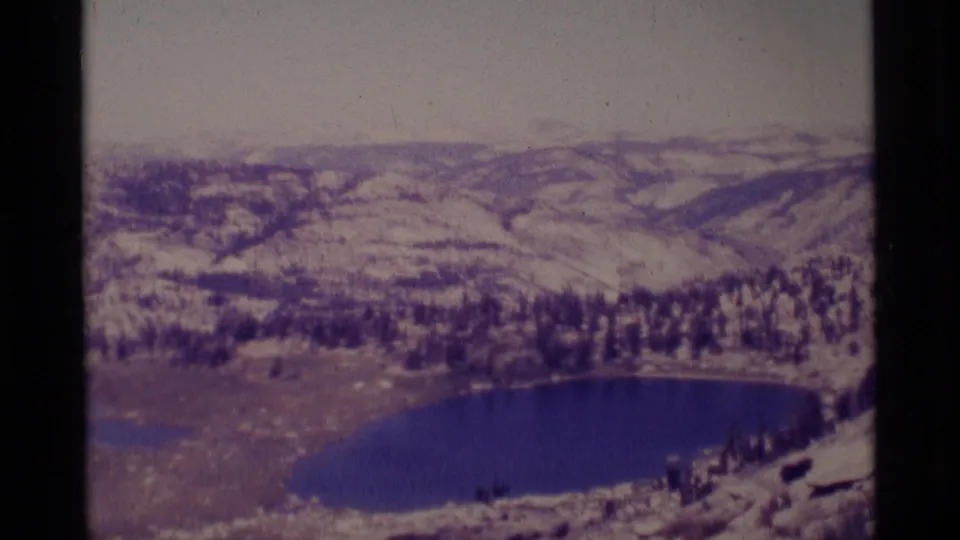1975 8mm view of a Minaret Wilderness lake surrounded by evergreen trees, pale slopes, and distant ridgelines.