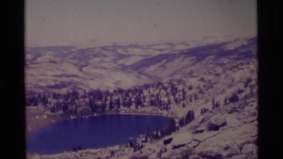 1975 8mm frame showing the lake edge, evergreen forest, rounded rocks, and snow-dusted Minaret Wilderness hills.