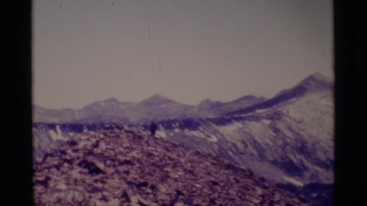 Wide rocky ridgeline with a small hiker and mountain peaks beyond.