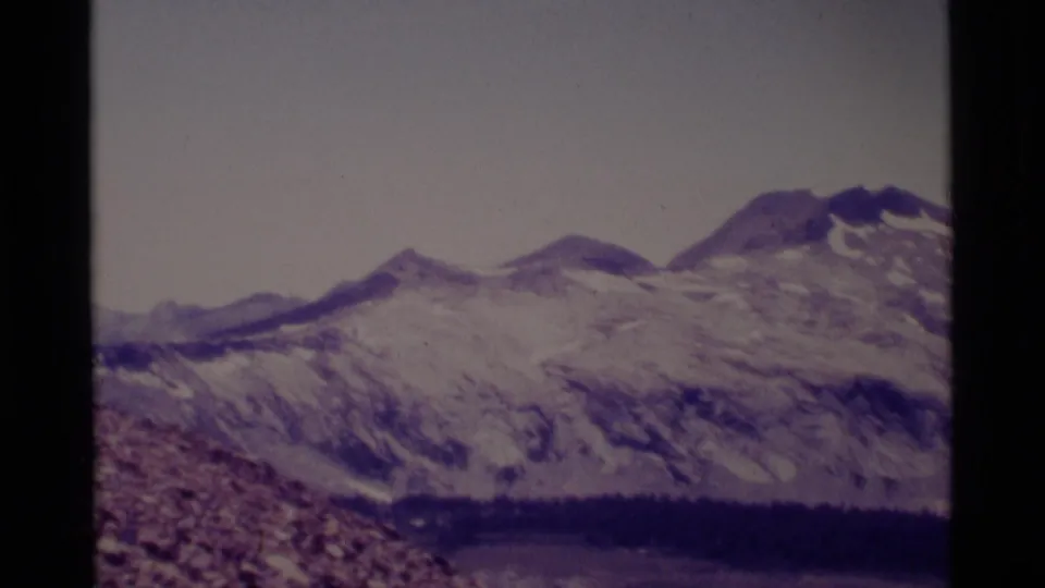 1975 8mm landscape frame showing Minaret Wilderness peaks, rocky slopes, snow patches, and a dark basin below.