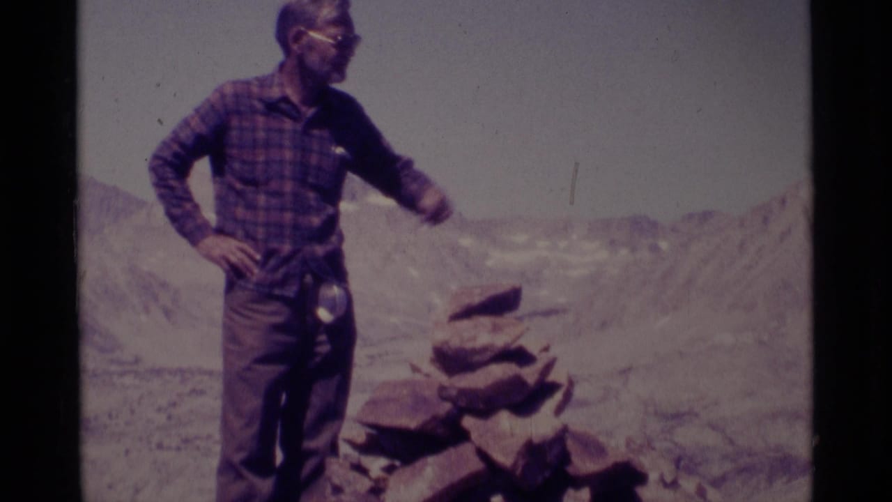 Adult man in a plaid shirt and sunglasses stands on rocky high ground in the Minaret Wilderness, California, with alpine ridges behind him.