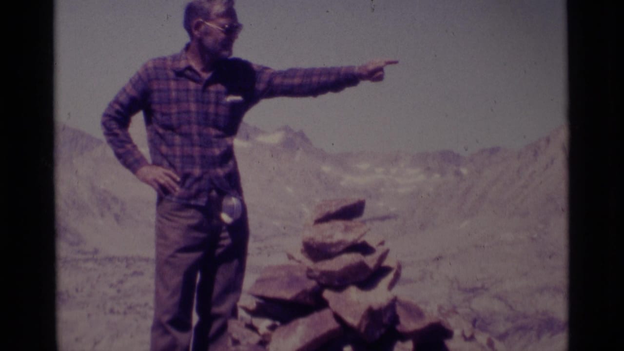 Man in plaid shirt points across a rocky mountain landscape.