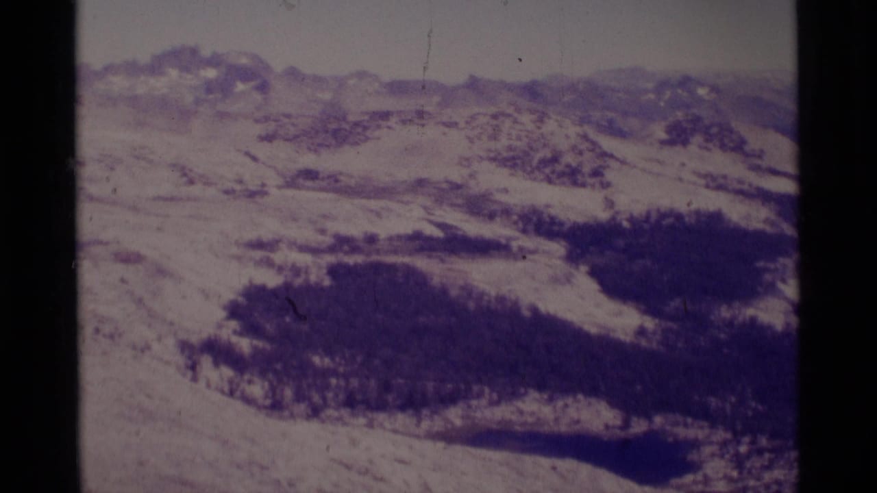 1975 8mm aerial view over Minaret Wilderness, showing snowy Sierra Nevada ridges, forested slopes, and a small dark lake.