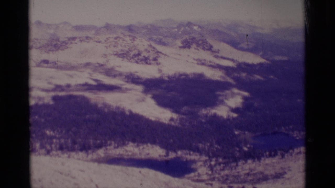 Aerial 8mm frame over Minaret Wilderness with snowfields, wooded basin areas, distant mountain layers, and small dark lakes.