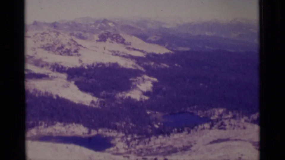 1975 aerial home-movie frame showing a broad snowy mountain basin in Minaret Wilderness with evergreen forest and dark alpine lakes.