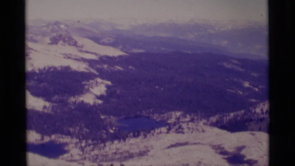 Vintage 8mm aerial view of Minaret Wilderness, with snowy Sierra Nevada slopes, forested valley terrain, and small alpine lakes.