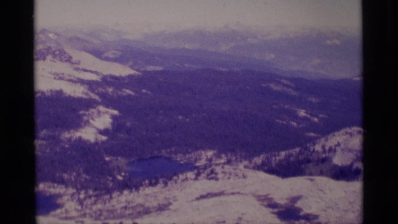 1975 8mm view from high in Minaret Wilderness, showing snowy mountain terrain, forested valley, and a small lake below.