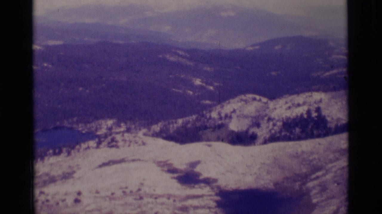 1975 8mm mountain panorama in Minaret Wilderness, with snow patches across ridges and hazy forested terrain beyond.