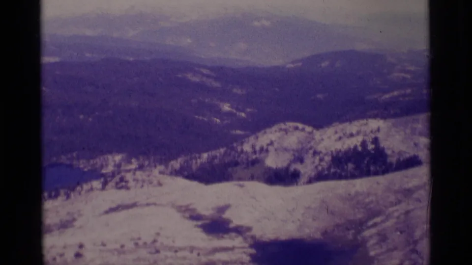 1975 8mm view across Minaret Wilderness terrain, with snow in the foreground, wooded slopes, and a small lake below.