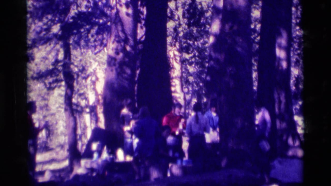 1975 8mm frame shows adults in a forested Minaret Wilderness campsite area, some near gear or a possible communal table.